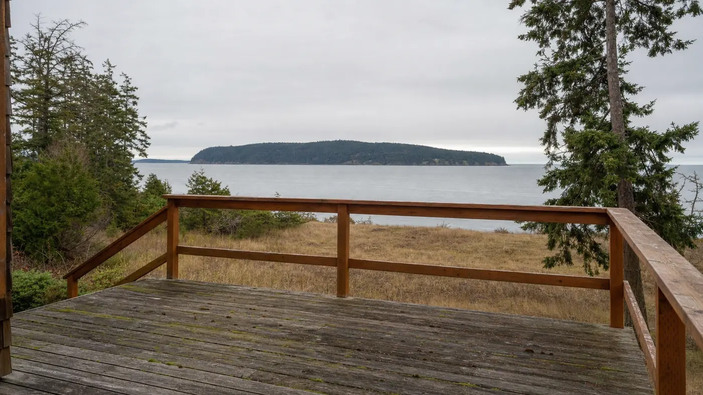 A weathered deck with a cedar railing looking out over calm gray water toward a forested island