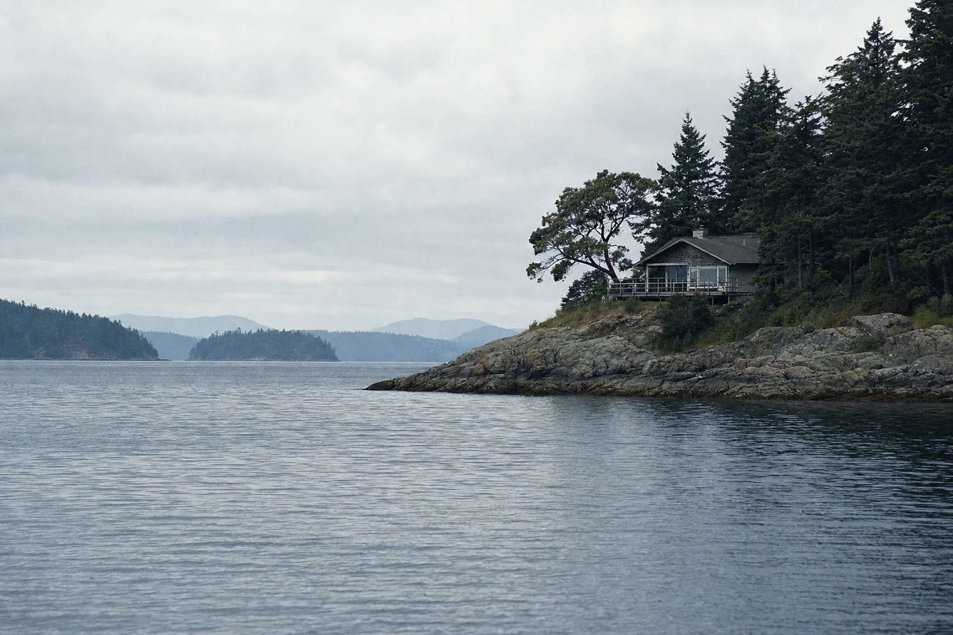 A cedar-shingled island home on a rocky point above calm water, seen from a boat on an overcast San Juan Islands morning